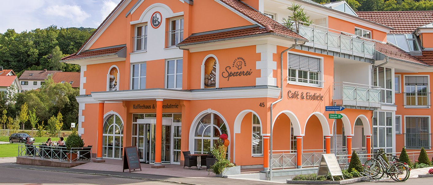 Orangefarbenes Gebäude mit Aufschrift 'Kaffeehaus-Spezerei Sanct Bernhard'. Menschen sitzen draußen an Tischen. Fahrräder stehen vor dem Gebäude., © Kräuterhaus Sanct Bernhard KG