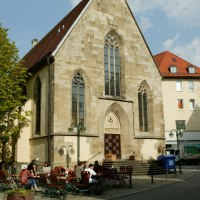 Die Nikolaikirche mit gotischen Fenstern, umgeben von Bäumen und Sitzgelegenheiten. Menschen sitzen draußen bei sonnigem Wetter., © Sphäre Verlag Die Nikolaikirche mit gotischen Fenstern, umgeben von Bäumen und Sitzgelegenheiten. Menschen sitzen draußen bei sonnigem Wetter., © Sphäre Verlag