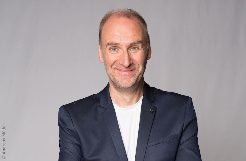 Ein Mann in einem dunklen Anzug mit weißem T-Shirt lächelt vor einem grauen Hintergrund., © Verein der Freunde des Lions Club Kornwestheim e.V.