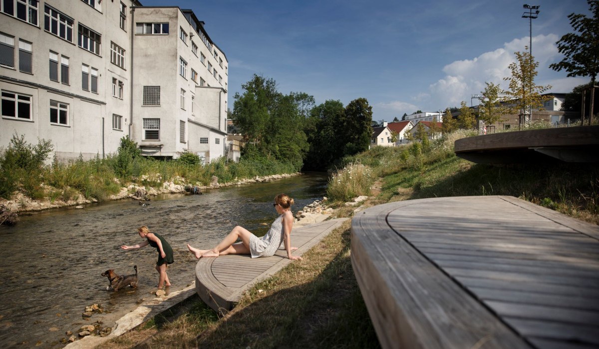 Zwei Frauen entspannen an einem Fluss neben einem alten Industriegebäude. Eine Frau spielt mit einem Hund im Wasser, während die andere auf einer Holzplattform sitzt., © Stoppel