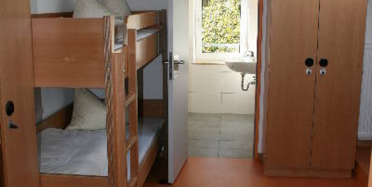 Ein einfach eingerichtetes Zimmer in einer Jugendherberge mit Etagenbett, Einzelbett, Schrank und Fenster. Helle Holzmöbel dominieren den Raum.