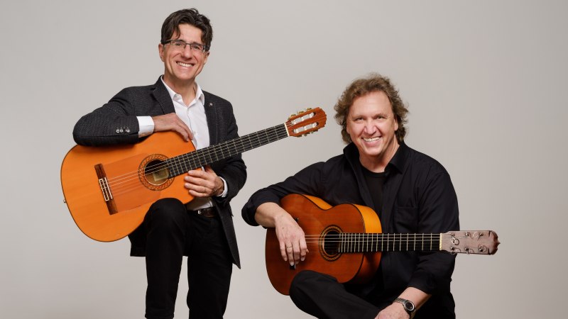 Zwei M&auml;nner posieren mit Gitarren und l&auml;cheln in die Kamera. Der eine steht, der andere sitzt. Beide tragen elegante Kleidung.