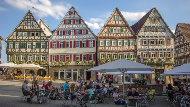 Fachwerkhäuser am Marktplatz in Herrenberg, Menschen sitzen an Tischen im Freien unter Sonnenschirmen, blauer Himmel im Hintergrund., © Natur.Nah. Schönbuch & Heckengäu
