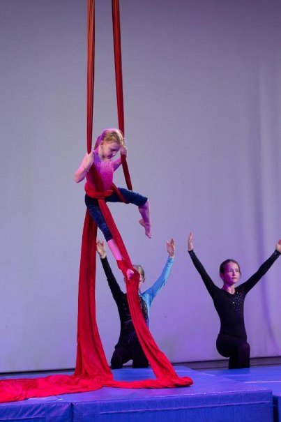 Ein Kind übt Luftakrobatik an roten Tüchern, zwei andere Kinder knien auf einer Matte und heben die Arme., © Kunstschule Labyrinth