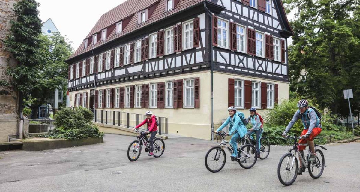 Vier Radfahrer fahren an einem historischen Fachwerkhaus in Kirchheim unter Teck vorbei. Das Gebäude hat weiße Wände und braune Fensterläden., © Schwäbische Alb Tourismusverband e.V.