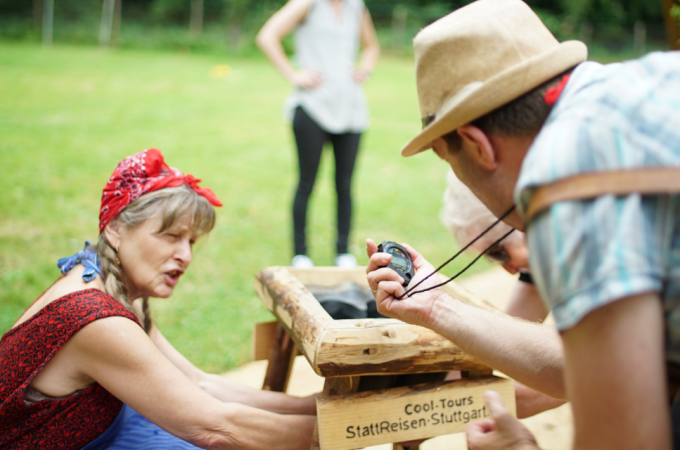 Eine Frau und ein Mann bei einer Outdoor-Aktivität. Der Mann hält eine Stoppuhr, die Frau arbeitet an einer Holzkonstruktion. Im Hintergrund eine weitere Person., © Cool-Tours StattReisen Eine Frau und ein Mann bei einer Outdoor-Aktivität. Der Mann hält eine Stoppuhr, die Frau arbeitet an einer Holzkonstruktion. Im Hintergrund eine weitere Person., © Cool-Tours StattReisen
