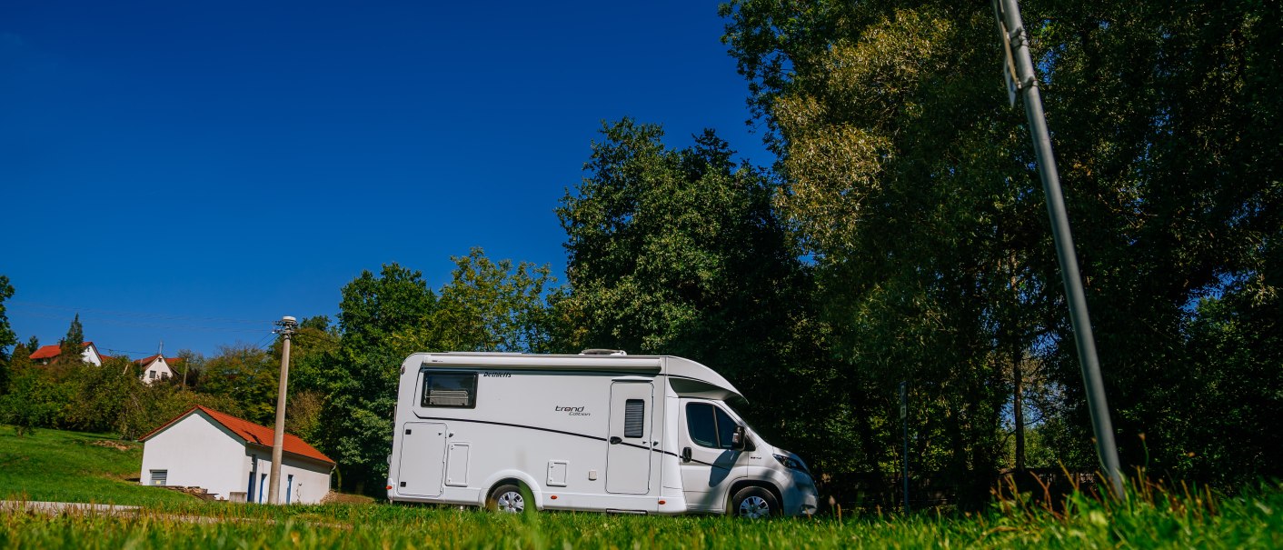 Ein Wohnmobil steht auf einem grünen Stellplatz, umgeben von Bäumen und einem klaren blauen Himmel., © SMG, Thomas Niedermüller