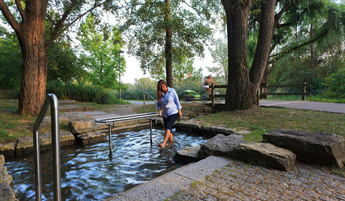 Eine Frau genießt eine Kneippanlage im Freien, umgeben von Bäumen und Natur. Ein Radfahrer ist im Hintergrund zu sehen., © Remstal Tourismus e.V.