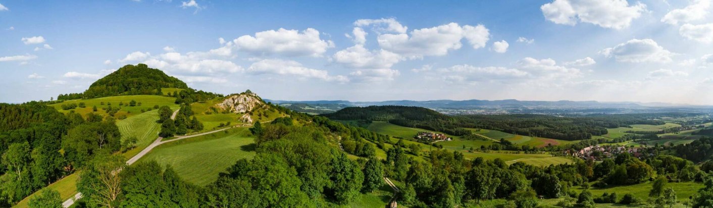 Grüne Hügellandschaft mit Bäumen und Feldern unter blauem Himmel mit weißen Wolken. Im Hintergrund sind Dörfer und Wälder zu sehen. Grüne Hügellandschaft mit Bäumen und Feldern unter blauem Himmel mit weißen Wolken. Im Hintergrund sind Dörfer und Wälder zu sehen.