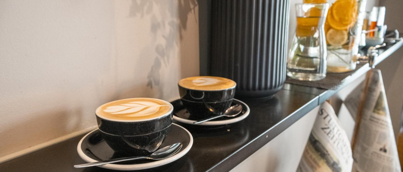 Zwei Cappuccino-Tassen auf einem Regal, daneben Karaffen mit Zitrusfrüchten und Zeitungen. Gemütliche Café-Atmosphäre., © SMG, Sarah Schmid Zwei Cappuccino-Tassen auf einem Regal, daneben Karaffen mit Zitrusfrüchten und Zeitungen. Gemütliche Café-Atmosphäre., © SMG, Sarah Schmid
