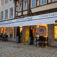 Außenansicht eines Restaurants mit Café und Konditorei. Beleuchtete Fenster und Tische im Freien bei Dämmerung. Schriftzug 'AlbThermen am Markt' sichtbar.
