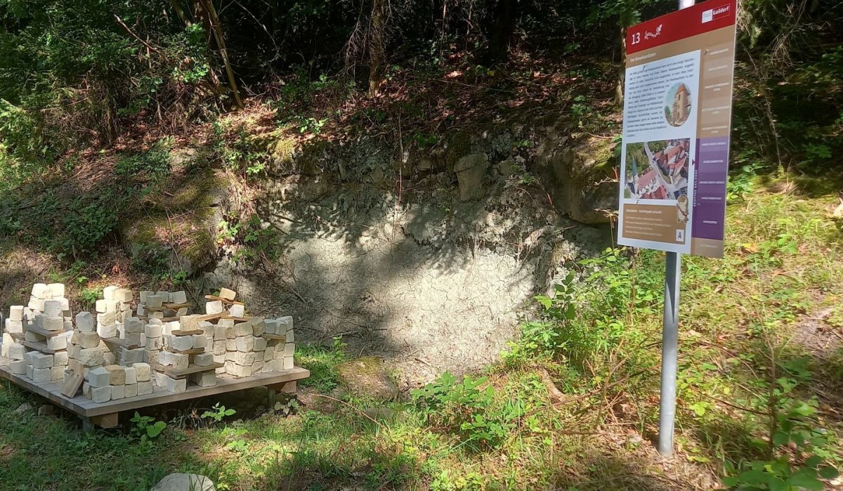 Geopfad Station 13 in Gaildorf: Eine Infotafel steht neben einem Haufen heller Steine auf einer Holzpalette, umgeben von Wald.