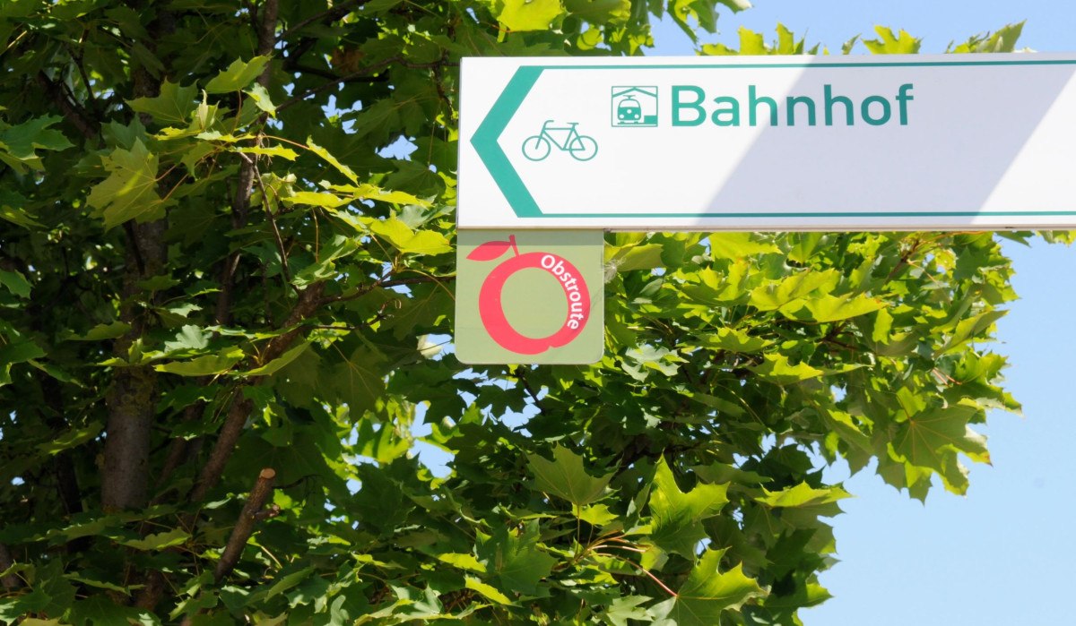 Ein Wegweiser zeigt zur Obstroute und zum Bahnhof, umgeben von grünen Blättern., © Baumgärtner Ein Wegweiser zeigt zur Obstroute und zum Bahnhof, umgeben von grünen Blättern., © Baumgärtner