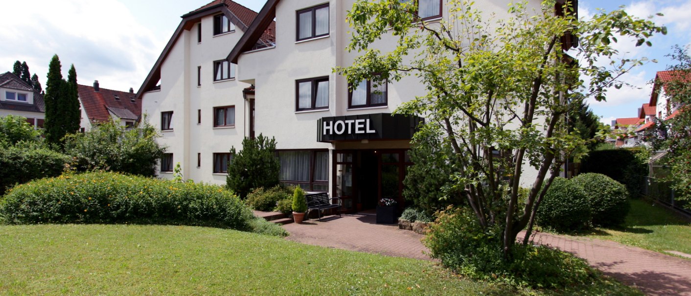 Ein Hotelgebäude mit weißer Fassade und einem Schild "HOTEL" über dem Eingang, umgeben von gepflegtem Garten und Bäumen., © Hotel Flora
