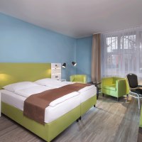 Modernes Doppelzimmer mit grünem Bett, Schreibtisch, zwei Sesseln und TV. Helle Wände und große Fenster sorgen für eine freundliche Atmosphäre., © TOMAS