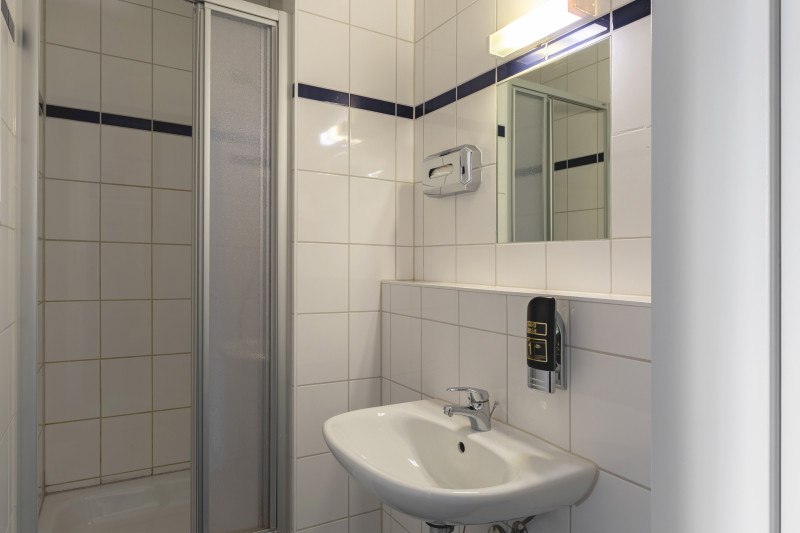 Kleines, gefliestes Badezimmer mit Dusche, Waschbecken und Spiegel. Ein Seifenspender ist an der Wand montiert., © A&O Hostels Marketing GmbH