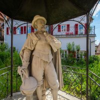 Steinstatue eines Mannes mit Hut und Umhang, der ein Instrument hält. Sie steht unter einem Pavillon in einem grünen Garten, umgeben von Häusern., © Stuttgart-Marketing GmbH, Achim Mende Steinstatue eines Mannes mit Hut und Umhang, der ein Instrument hält. Sie steht unter einem Pavillon in einem grünen Garten, umgeben von Häusern., © Stuttgart-Marketing GmbH, Achim Mende