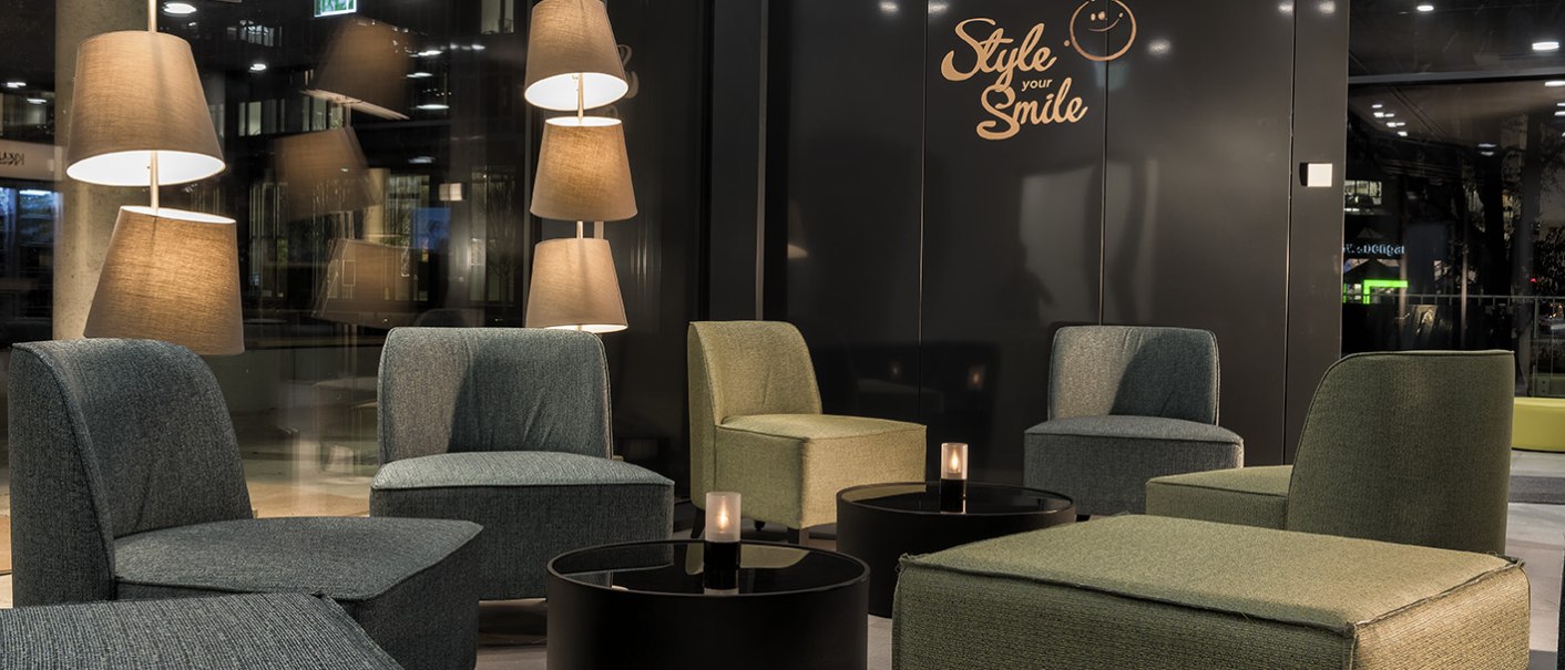 Moderne Lobby mit bequemen Sesseln, runden Tischen und stilvollen Stehlampen. An der Wand steht "Style your Smile". Gemütliche Atmosphäre., © ibis Styles Stuttgart Vaihingen