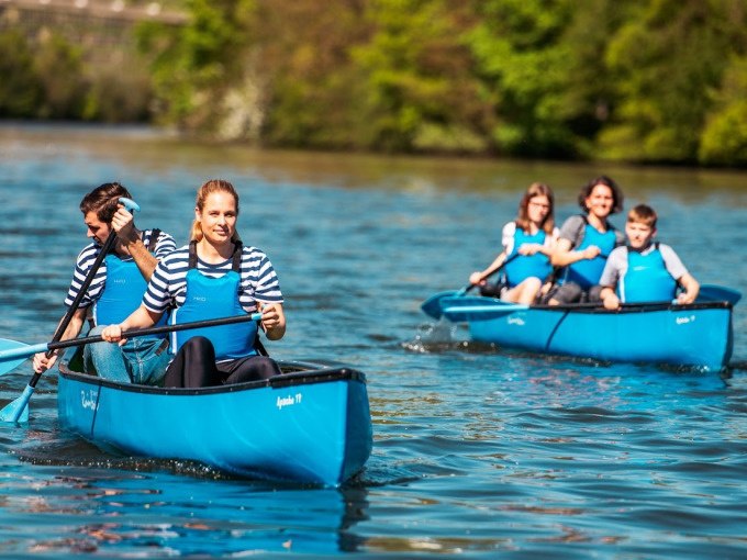 Zwei blaue Kanus mit Menschen paddeln auf einem Fluss. Die Personen tragen gestreifte Shirts und blaue Schwimmwesten. Im Hintergrund ist grüne Natur zu sehen., © Cool-Tours StattReisen Zwei blaue Kanus mit Menschen paddeln auf einem Fluss. Die Personen tragen gestreifte Shirts und blaue Schwimmwesten. Im Hintergrund ist grüne Natur zu sehen., © Cool-Tours StattReisen