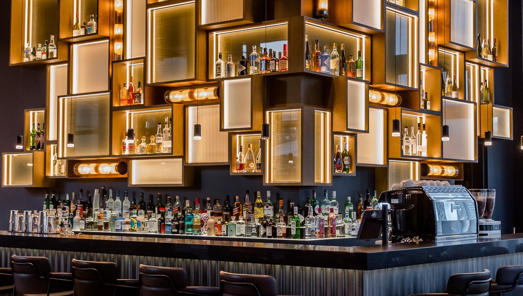 Eine stilvolle Bar mit beleuchteten Regalen voller Flaschen, modernen Barhockern und einer eleganten Theke., © TOMAS Eine stilvolle Bar mit beleuchteten Regalen voller Flaschen, modernen Barhockern und einer eleganten Theke., © TOMAS