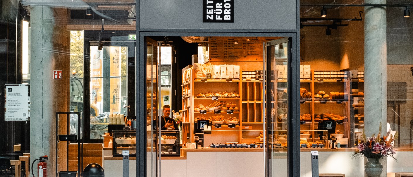 Eingang einer Bäckerei mit Glasfront. Im Inneren sind Regale mit Backwaren zu sehen. Über der Tür steht 'Zeit für Brot'., © Zeit für Brot Stuttgart
