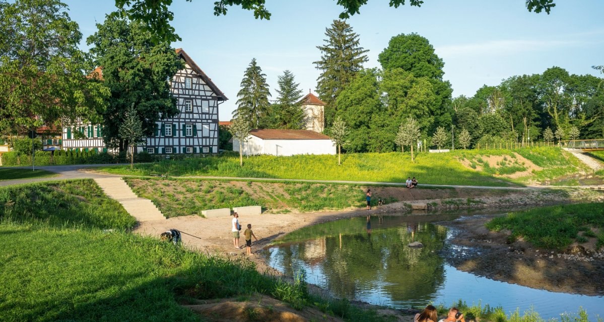 Fachwerkhaus neben einem kleinen Fluss, umgeben von gr&uuml;ner Landschaft. Menschen entspannen am Ufer unter blauem Himmel., &copy; Remstal Tourismus e.V.