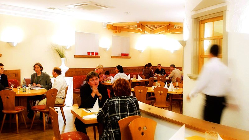 Gemütliches Restaurant mit Holzmöbeln, mehreren Gästen an Tischen und einem Kellner in Bewegung. Warme Beleuchtung und dekorative Elemente an den Wänden., © SMG