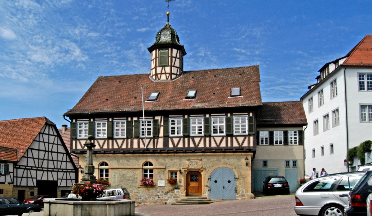 Historisches Fachwerkgebäude mit kleinem Turm und Brunnen im Vordergrund. Autos parken auf dem Platz, blauer Himmel im Hintergrund., © Natur.Nah. Schönbuch & Heckengäu Historisches Fachwerkgebäude mit kleinem Turm und Brunnen im Vordergrund. Autos parken auf dem Platz, blauer Himmel im Hintergrund., © Natur.Nah. Schönbuch & Heckengäu
