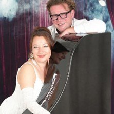 Ein Mann und eine Frau lächeln an einem schwarzen Flügel. Der Hintergrund ist bühnenartig mit Vorhängen und Lichtern gestaltet., © links im Bild Ein Mann und eine Frau lächeln an einem schwarzen Flügel. Der Hintergrund ist bühnenartig mit Vorhängen und Lichtern gestaltet., © links im Bild