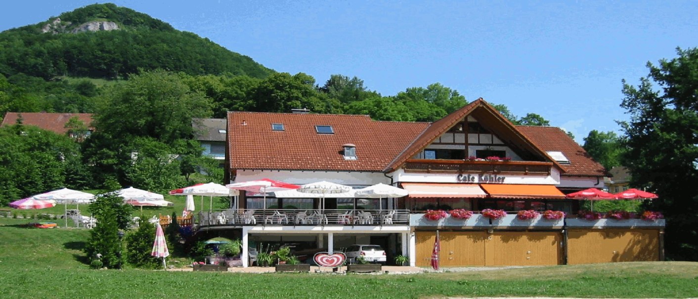 Café Köhler mit Terrasse und Sonnenschirmen, umgeben von grüner Landschaft und einem bewaldeten Hügel im Hintergrund., © Café Köhler