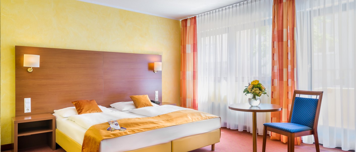 Ein gemütliches Hotelzimmer mit Doppelbett, gelben Wänden und orangefarbenen Vorhängen. Ein Tisch mit Blumen und ein blauer Stuhl ergänzen das Ambiente., © TOMAS