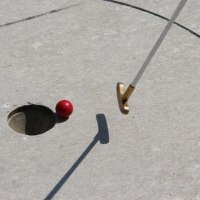 Ein roter Minigolfball liegt knapp neben einem Loch auf einer Betonfläche. Ein goldener Schläger ist bereit zum Schlag.