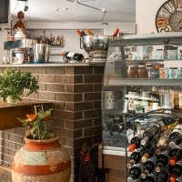 Gemütliches Café mit Holztisch, Weinregal, dekorativen Bildern und Pflanzen. Eine Theke mit Getränken und ein Fernseher sind ebenfalls zu sehen., © SMG, Sarah Schmid Gemütliches Café mit Holztisch, Weinregal, dekorativen Bildern und Pflanzen. Eine Theke mit Getränken und ein Fernseher sind ebenfalls zu sehen., © SMG, Sarah Schmid