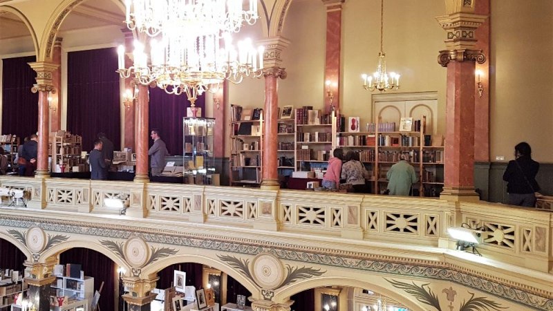 Blick in die Musikhalle Ludwigsburg mit antiken Bücherständen auf zwei Etagen, dekorativen Säulen und Kronleuchtern. Besucher stöbern in Büchern., © Petra Bewer - Architektur-Antiquariat