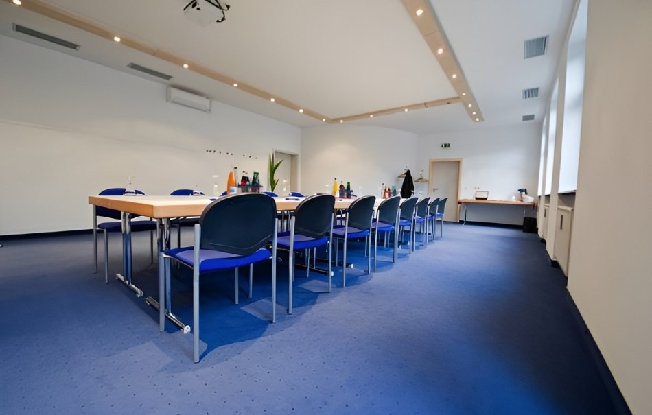 Leerer Konferenzraum mit großem Tisch, blauen Stühlen und blauem Teppichboden. An der Wand sind Getränke und ein Projektor zu sehen., © Hotel Neuwirtshaus Leerer Konferenzraum mit großem Tisch, blauen Stühlen und blauem Teppichboden. An der Wand sind Getränke und ein Projektor zu sehen., © Hotel Neuwirtshaus