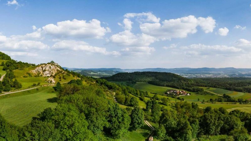 Grüne Hügellandschaft mit Bäumen und Feldern unter blauem Himmel mit weißen Wolken. Im Hintergrund sind Dörfer und Wälder zu sehen. Grüne Hügellandschaft mit Bäumen und Feldern unter blauem Himmel mit weißen Wolken. Im Hintergrund sind Dörfer und Wälder zu sehen.