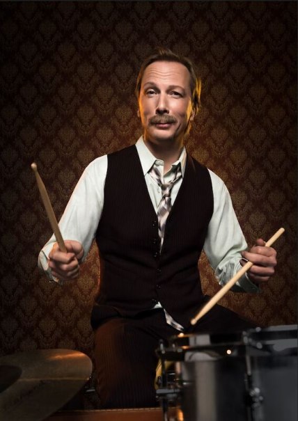 Ein Mann in Weste und Hemd spielt Schlagzeug vor einer gemusterten Tapete. Er hält zwei Drumsticks in den Händen., © C² CONCERTS GmbH