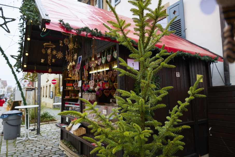 Ein Weihnachtsmarktstand mit roten Dach und Dekorationen. Lebkuchenherzen hängen aus. Ein Tannenzweig im Vordergrund., © SMG, Sarah Schmid