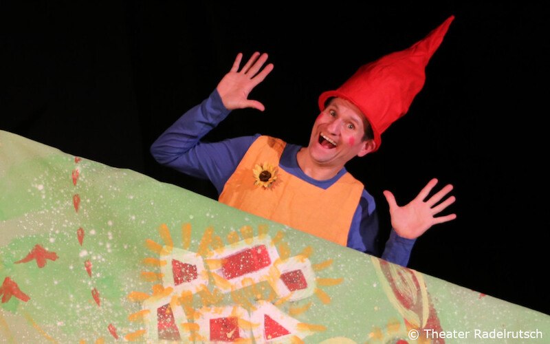 Ein Schauspieler in einem Zwergenkostüm mit roter Mütze und orangefarbenem Oberteil lächelt und hebt die Hände vor einem bunten Hintergrund., © Kultur- und Sportamt Stadt Bietigheim-Bissingen