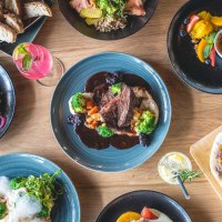 Ein Tisch mit verschiedenen Gerichten: Fleisch mit Gemüse, Salate, Burger mit Pommes, Brot und Desserts. Getränke sind ebenfalls zu sehen., © SatzDrei