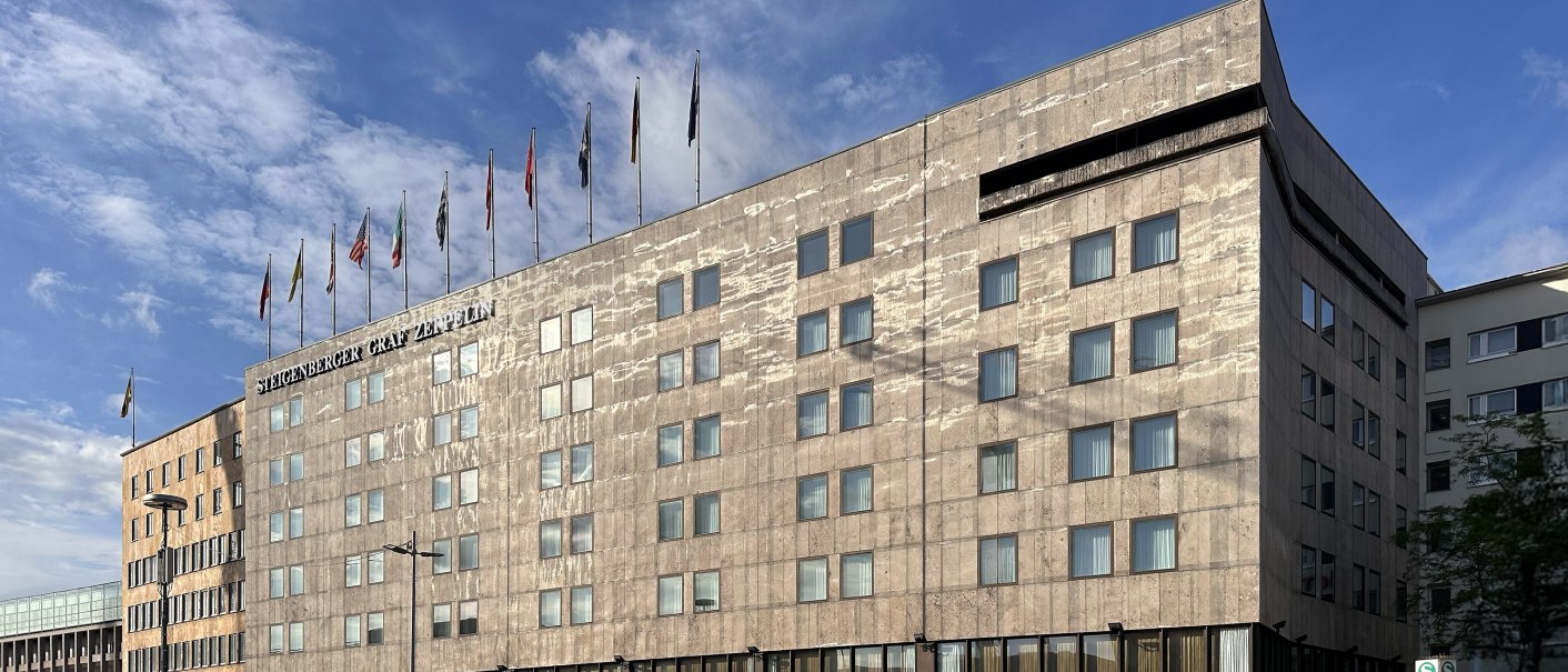 Das Steigenberger Graf Zeppelin Hotel mit Flaggen auf dem Dach vor blauem Himmel. Moderne Architektur mit vielen Fenstern., © Steigenberger Graf Zeppelin