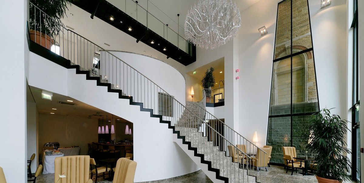 Elegante Lobby mit geschwungener Treppe, gro&szlig;em Kronleuchter und gem&uuml;tlichem Sitzbereich. Hohe Fenster und Pflanzen schaffen eine einladende Atmosph&auml;re., &copy; Arcotel Hotels