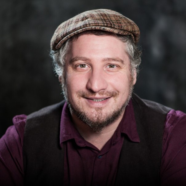 Ein Mann mit Schiebermütze und Bart lächelt in die Kamera. Er trägt ein lila Hemd und eine dunkle Weste. Der Hintergrund ist dunkel und unscharf., © Mauerwerk GmbH Ein Mann mit Schiebermütze und Bart lächelt in die Kamera. Er trägt ein lila Hemd und eine dunkle Weste. Der Hintergrund ist dunkel und unscharf., © Mauerwerk GmbH