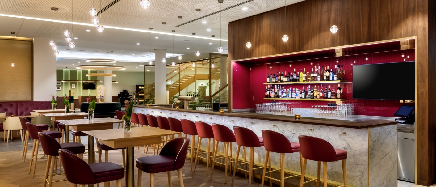 Moderne Bar mit roten Stühlen, Marmor-Theke und beleuchtetem Regal mit Spirituosen. Helle Deckenbeleuchtung und Holzboden schaffen eine einladende Atmosphäre., © Mövenpick Hotel Stuttgart Messe & Congress Moderne Bar mit roten Stühlen, Marmor-Theke und beleuchtetem Regal mit Spirituosen. Helle Deckenbeleuchtung und Holzboden schaffen eine einladende Atmosphäre., © Mövenpick Hotel Stuttgart Messe & Congress