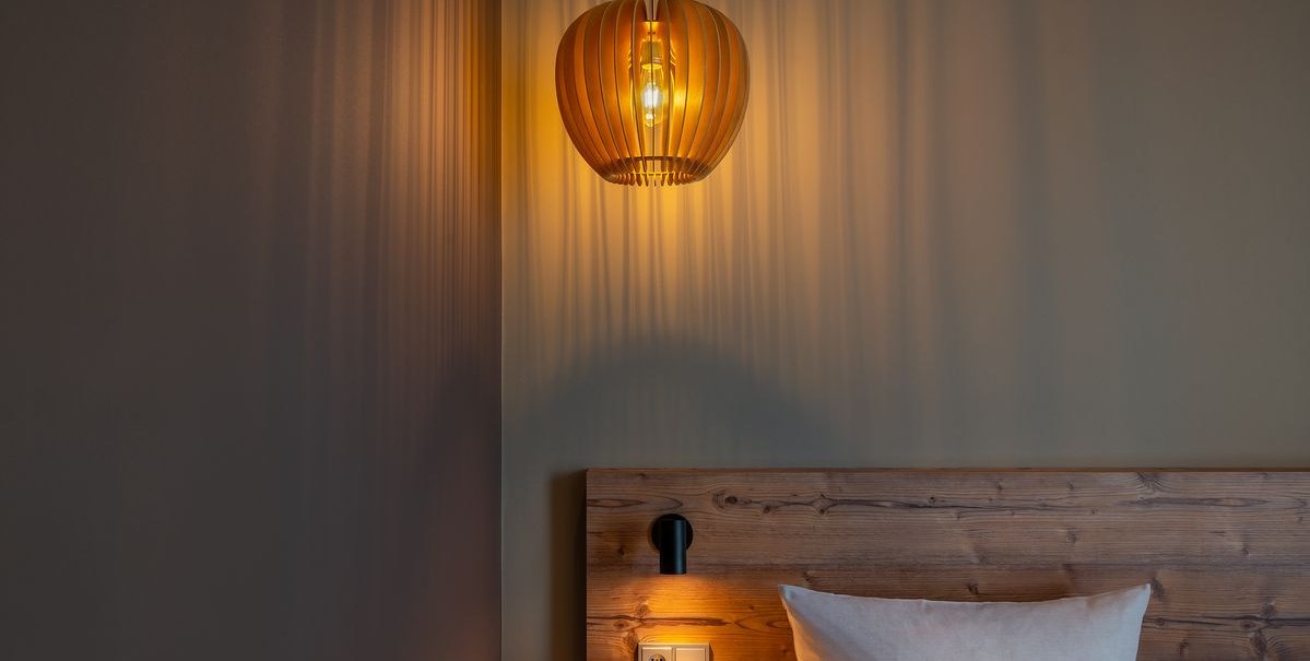 Eine hölzerne Lampe wirft warmes Licht auf eine Wand und ein Holzbett mit Kissen in einem modernen Zimmer., © TOMAS Eine hölzerne Lampe wirft warmes Licht auf eine Wand und ein Holzbett mit Kissen in einem modernen Zimmer., © TOMAS