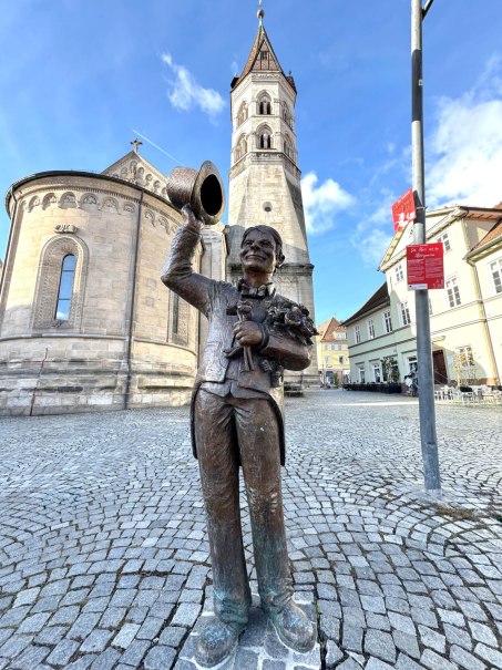 Zu sehen ist eine Bronzefigur, die den Gmünder "Alois" darstellt. Im Hintergrund sieht man die Johanniskirche., © Touristik und Marketing GmbH Zu sehen ist eine Bronzefigur, die den Gmünder "Alois" darstellt. Im Hintergrund sieht man die Johanniskirche., © Touristik und Marketing GmbH