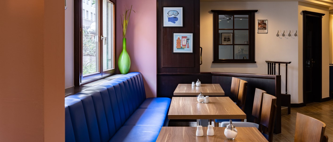 Ein einladender Fr&uuml;hst&uuml;cksraum mit blauen Sitzb&auml;nken, Holztischen und dekorativen Bildern an den W&auml;nden. Gro&szlig;e Fenster lassen Tageslicht herein., &copy; Novum  Hospitality