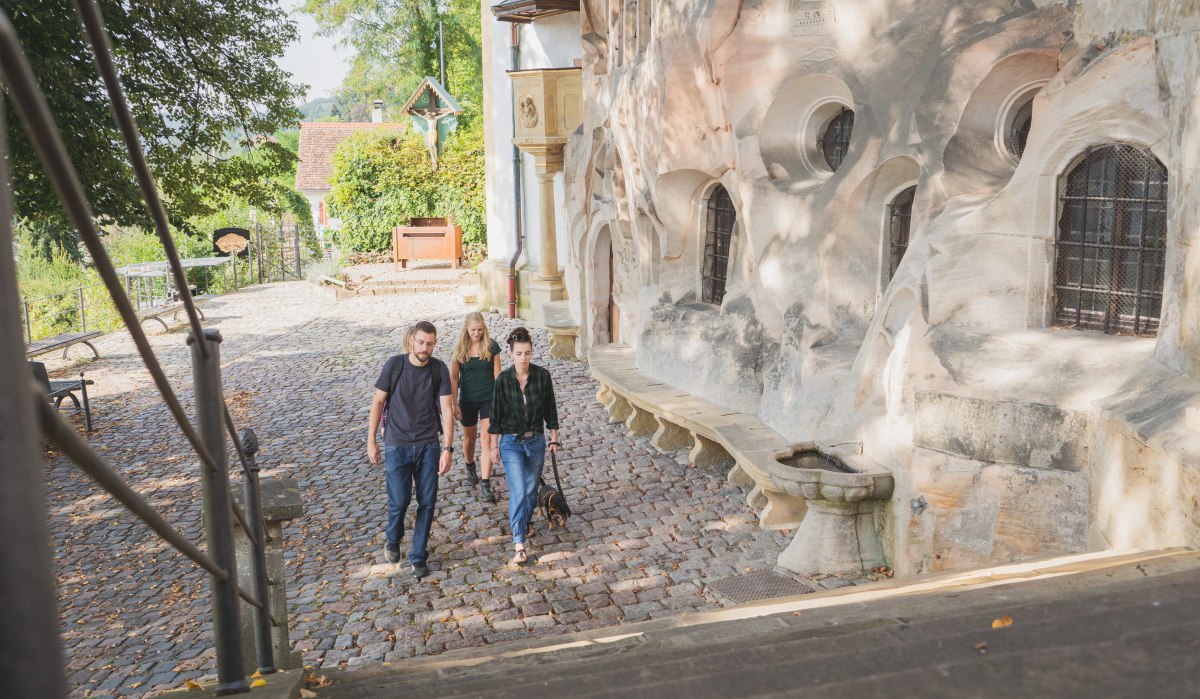 Vier Personen spazieren auf einem gepflasterten Weg neben einer historischen Steinmauer in Schw&auml;bisch Gm&uuml;nd. Im Hintergrund sind B&auml;ume und ein kleines Geb&auml;ude zu sehen.