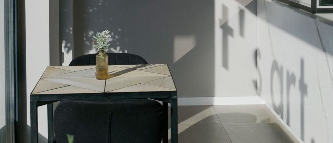 Heller Raum mit Tisch, zwei schwarzen St&uuml;hlen und einer kleinen Vase; an der Wand f&auml;llt der Schatten des Wortes &bdquo;Short&ldquo;., &copy; Taraba