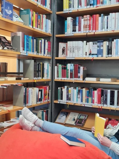 Eine Person liegt entspannt auf einem roten Sitzsack in einer Bibliothek und liest ein gelbes Buch. Umgeben von Bücherregalen voller Bücher., © (c) Regina Gobert Eine Person liegt entspannt auf einem roten Sitzsack in einer Bibliothek und liest ein gelbes Buch. Umgeben von Bücherregalen voller Bücher., © (c) Regina Gobert